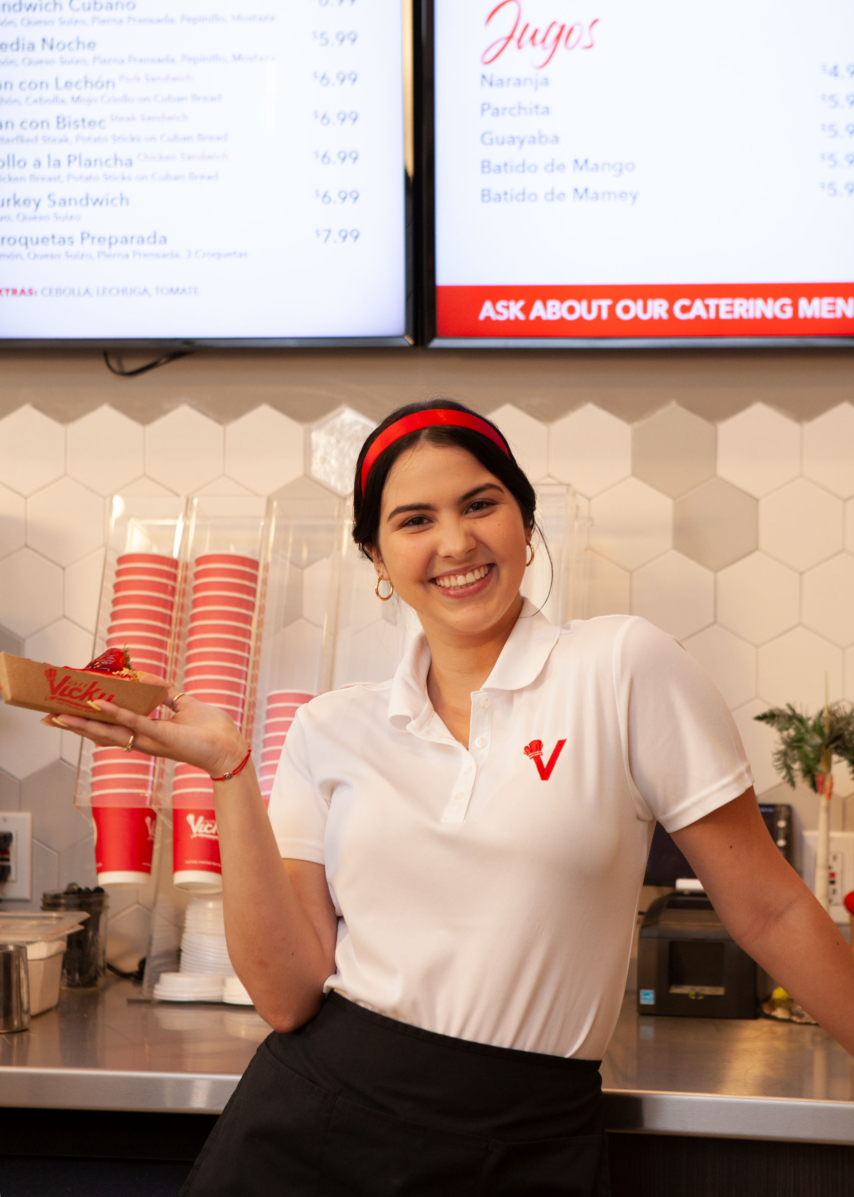 A Vicky Bakery team member behind the West Pines counter, holding a fresh order in front of the Cuban menu.